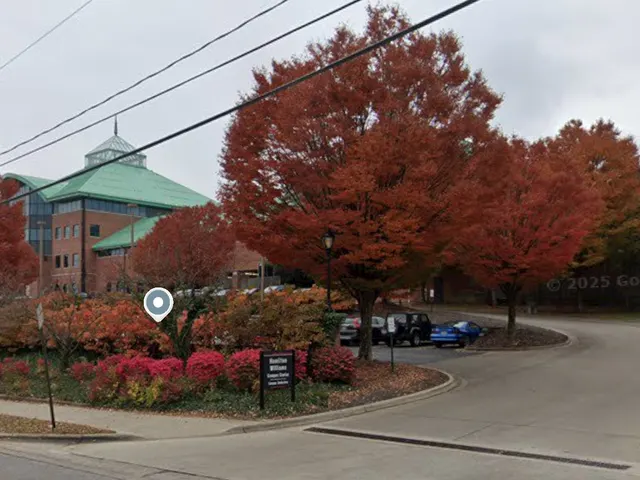 Street view from Spring St: the Hamilton-Williams Campus Center sign at the drive into its parking lot, with the building behind the trees.